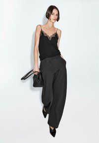 Camisole noire avec dentelle, associée à un pantalon noir ample. Le modèle tient un petit sac à main noir et porte des escarpins noirs à bout pointu. Fond neutre.