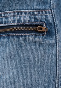 Artimas vaizdas į dalinai atidarytą žalvarinį užtrauktuką ant šviesiai mėlyno džinso audinio su matomomis siuvimo detalėmis.