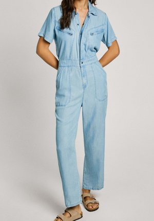 Light blue denim jumpsuit with short sleeves, front zip pockets, a fitted waist, and straight leg design, paired with brown sandals.