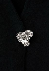 Broche floral plateada con un diseño de tres pétalos, textura pulida y detalles intrincados, situada sobre un fondo de tela negra.