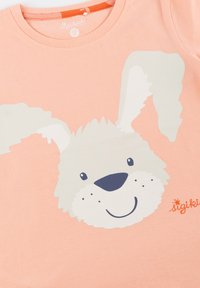 Peach cotton t-shirt featuring a large, light gray rabbit face graphic with floppy ears, black eyes, and a small nose. Brand name in orange.