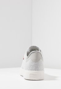 Vit stickad sneakers med grå hälflik, röd logotab på sidan och präglad Levi Strauss & Co. etikett på den vita sulan, sedd från baksidan.