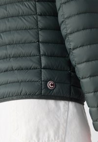 Mørkegrøn pufferjakke med quiltet tekstur, rund logo accente og en elastisk kant med brandingdetalje. Hvid denim synlig nedenfor.
