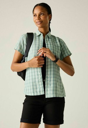 Donna che indossa una camicia a maniche corte a quadri verde, pantaloncini neri, zaino e smartwatch, in piedi con le mani giunte davanti a uno sfondo chiaro.