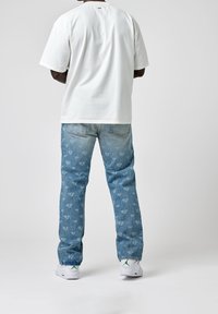 Vit bomullströja med avspänt snitt, parat med ljusblå denimjeans med ett mönster av logotyp över hela, samt fransar i kanten, bärs med vita sneakers.