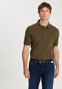 Oliwkowa koszulka polo z kołnierzykiem, zapięciem na dwa guziki i haftowanym logo; zestawiona z ciemnoniebieskimi dżinsami i czarnym paskiem.