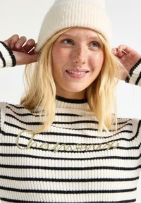 Strikket sweater med striber i creme og sort, med ribbede skuldre og manchetter; har guldskrift i kursiv "Champion" på forsiden. Strikket hue på hovedet.