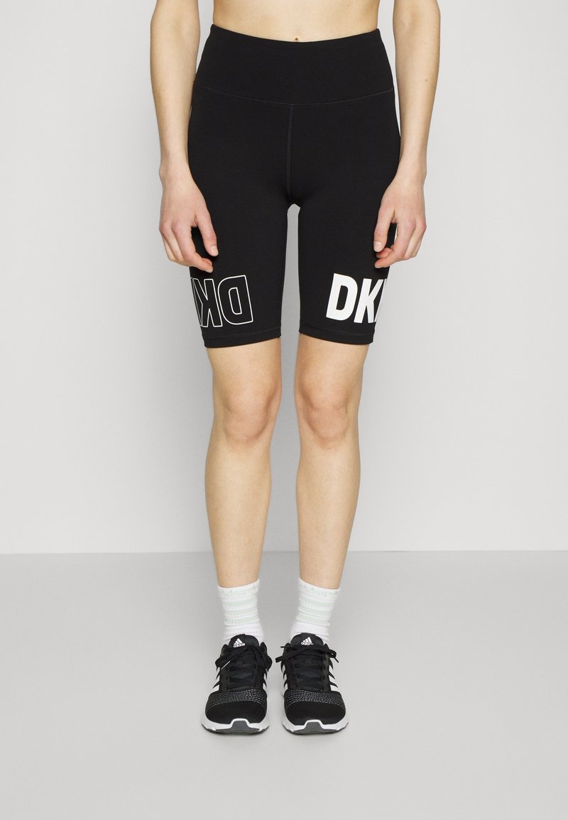 Personne portant des leggings noirs mi-genoux avec le texte "DKNY" sur les cuisses, des chaussettes blanches et des chaussures de course Adidas noires, debout sur un sol gris.