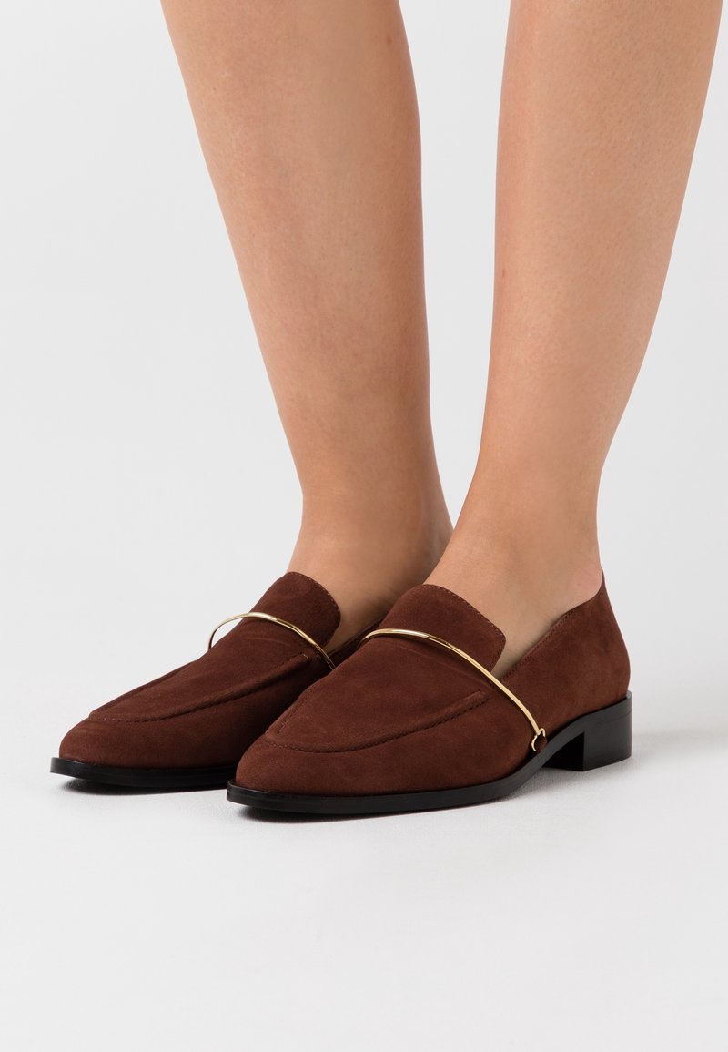 Mocassins en daim marron avec détail de bande métallique dorée, portés pieds nus, présentant des talons noirs bas et des bouts arrondis.
