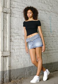 Top negru cu umeri descoperiți, fustă mini din denim de culoare albastră deschisă cu un curea largă și teniși albi, plasate pe un zid din cărămidă texturată.