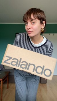 Kartono dėžė su pilku "zalando" logotipu, laikoma asmens su dryžuota marškinėliais. Fone matoma žalia siena ir rudi baldai.