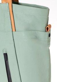 Groene draagtas met een gladde textuur, voorzien van een tan lederen handvat en een zijvak met knoopsluiting en een rits hoofdcompartiment.