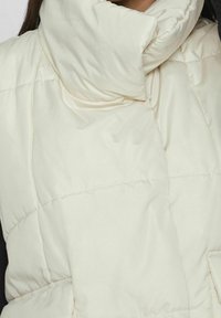 Puffig vit ärmlös jacka med hög krage, med kviltade sektioner och slät текстur. Accenter inkluderar en sidoficka och vikt tyg.