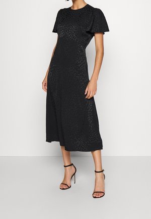 Vrouw draagt een zwarte midi-jurk met subtiele luipaardprint, vlindermouwen en zwarte hoge sandalen met enkelriempje, staand tegen een effen achtergrond.