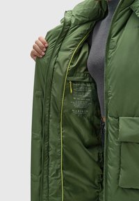 Zelena puf jakna s sjajnom, teksturiranom vanjštinom. Unutrašnjost sadrži svijetle žute naglaske, prostranu džep i detalje s tiskanim tekstom.