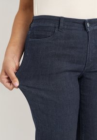 Närbild på en person som sträcker tyget av mörkblå denimjeans som bärs vid midjan tillsammans med en vit topp.