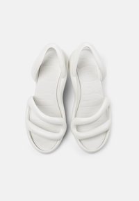 White sandals with a minimalist design, featuring wide, curved straps and a textured sole. Made of flexible material for comfort.