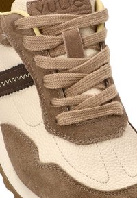 Zapatilla de color beige claro con acentos de ante marrón, cuero texturizado, puntera redonda y cordones a juego. Presenta una franja lateral marrón y un logo.