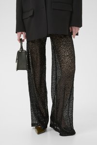 Pantaloni a gamba larga neri con paillettes, realizzati in un materiale texturizzato simile a pizzo. Abbinati a un blazer nero e a delle scarpe con tacco verdi, mentre si tiene in mano una borsa piccola.