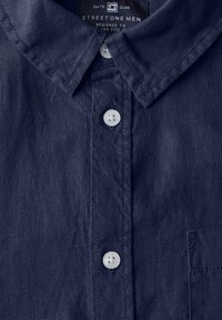 Námořnická modrá košile na knoflíky vyrobená z текстurované látky, disponující klasickým límečkem, malou kapsičkou a bílými knoflíky na légě.