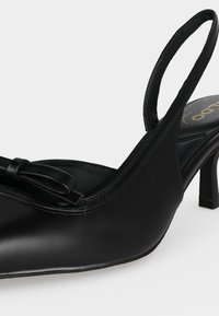 Escarpin slingback en cuir noir avec bout pointu et petit nœud décoratif sur le devant, finition lisse et talon fin de hauteur moyenne.