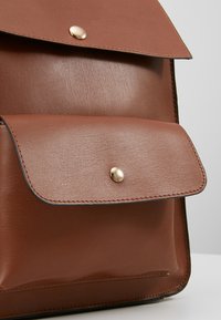 Ruskea nahkainen reppu, jossa on läpällinen yläosa, etutasku ja kultainen napsautuslukko. Sileä pinta ja pyöristetyt reunat.