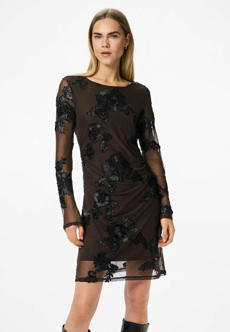 Juoda permatoma suknelė su ilgomis rankovėmis, papuošta juodais išsiuvinėtais gėlėmis ir surinkta liemens srityje. Suknelės ilgis iki vidurio šlaunies.