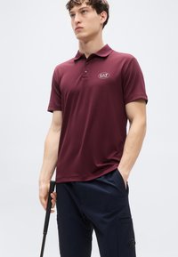 Μπλουζάκι πόλο σε μπορντό χρώμα, κατασκευασμένο από ελαφρύ ύφασμα, με γιακά, πάτο με τρία κουμπιά και λογότυπο EA7 στο στήθος. Συνδυάζεται με παντελόνια σε ναυτικό μπλε.