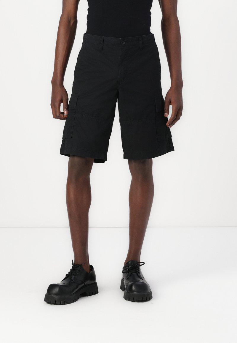 Pantalones cortos cargo negros con grandes bolsillos laterales, cierre de botón y un ajuste relajado, combinados con zapatos negros de suela gruesa.