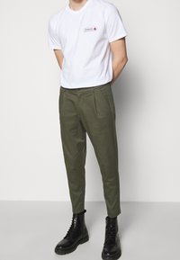 Bílé bavlněné tričko s červeným logem, kombinované s olivově zelen�ými kalhotami na míru s předními záhyby, nošené s černými šněrovacími botami.