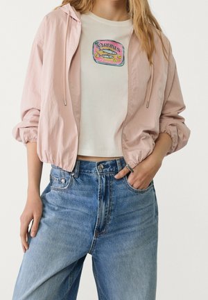 Vrouw draagt een lichtroze jas met rits over een wit cropped T-shirt met een afbeelding van een sardineblik, gecombineerd met high-waisted blauwe jeans.