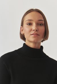 Maglione nero a collo alto con dettagli a coste e maniche raglan. Il modello ha i capelli corti e indossa orecchini a cerchio dorati, su uno sfondo chiaro.