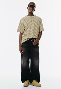 Beżowy, paskowany, oversize t-shirt, przechodzący w ciemny kolor na dole, w połączeniu z szerokimi czarnymi dżinsami i żółtymi butami roboczymi. Brązowa czapka na głowie.