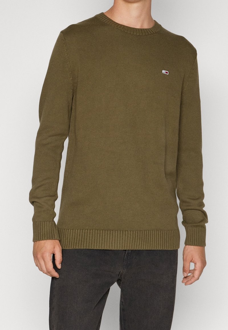 Homme portant un pull tricoté vert olive avec des poignets et un ourlet côtelés, associé à un jean noir.