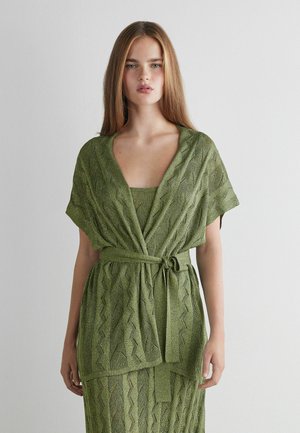 Jeune femme aux longs cheveux raides portant une robe portefeuille en maille verte avec une ceinture nouée, debout devant un fond blanc uni.