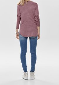 Langärmliges, strukturiertes bordeauxfarbiges Oberteil mit abgerundetem Saum, kombiniert mit enganliegenden blauen Jeans und weißen High-Top-Sneakern.