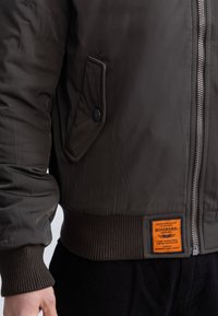 Kurtka bomberka z ciemnozielonego syntetycznego materia łu, z przednim zamkiem błyskawicznym, ściągaczami na rękawach i u dołu oraz wyraźną pomarańczową metką z boku.