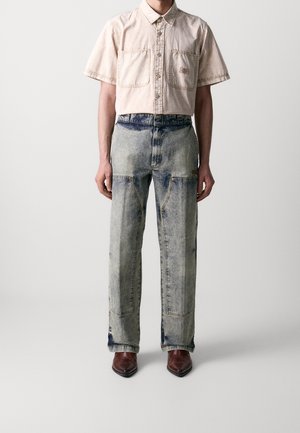 Persoon die een beige overhemd met korte mouwen draagt, een spijkerbroek met hoge taille en lichte wassing met voorzakken, en bruine, puntige schoenen.