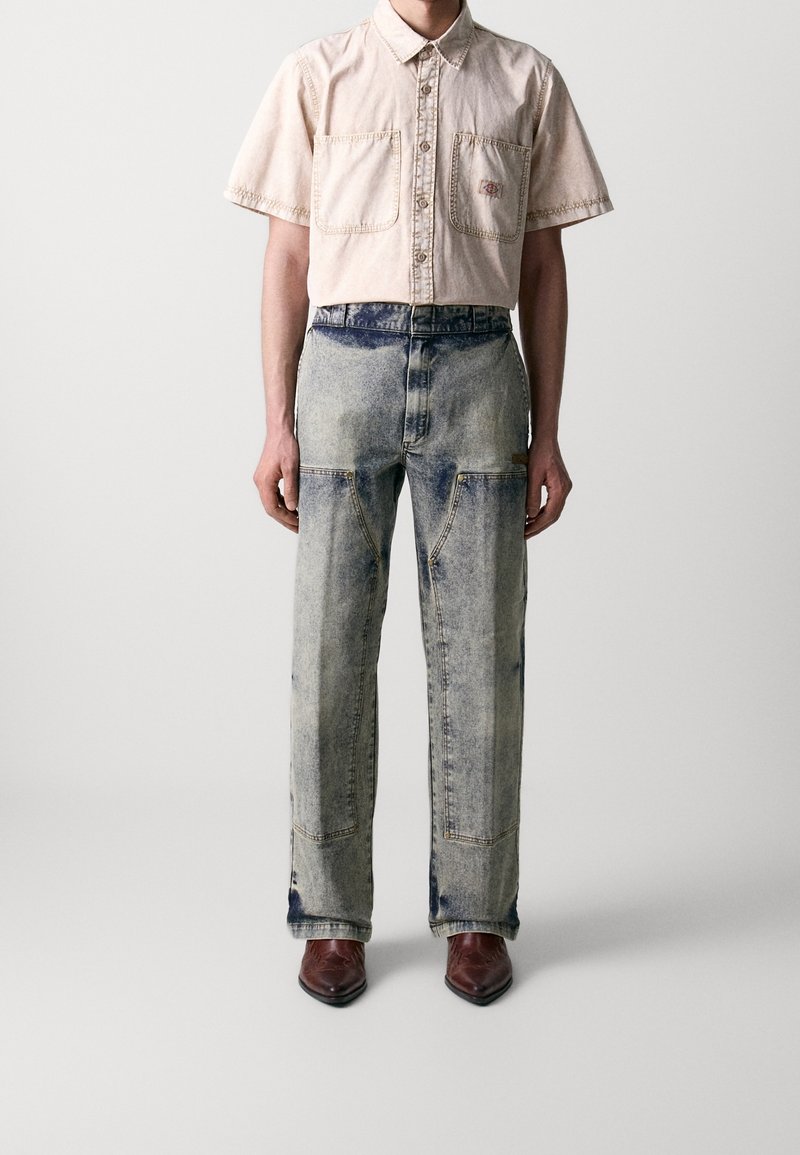 Personne portant une chemise beige à manches courtes avec boutons, un jean taille haute à effet délavé avec poches plaquées à l'avant, et des chaussures marron pointues.