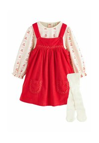 Red corduroy dress with straps and scalloped pockets, paired with a long-sleeve white top featuring cherry and bow prints, and white ribbed tights.