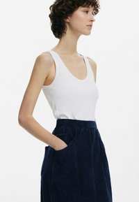 Kvinna som bär en vit ärmlös topp och en högmidjad marinblå kjol med fickor, stående i profil med en hand i fickan.