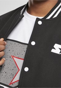 Zwarte varsityjas met witte gestreepte kraag en drukknoopsluiting, gedragen over een wit T-shirt met een grijze grafische print met een rode ster.