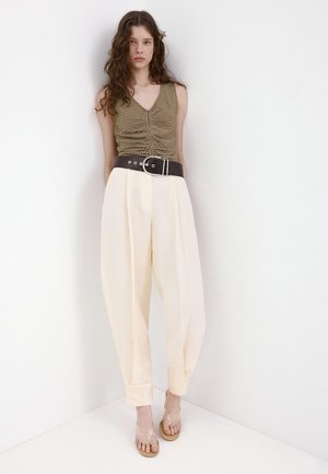 Femme debout contre un mur blanc, portant un top sans manches froncé de couleur olive, un pantalon plissé crème taille haute avec une ceinture noire et des talons à brides transparentes.