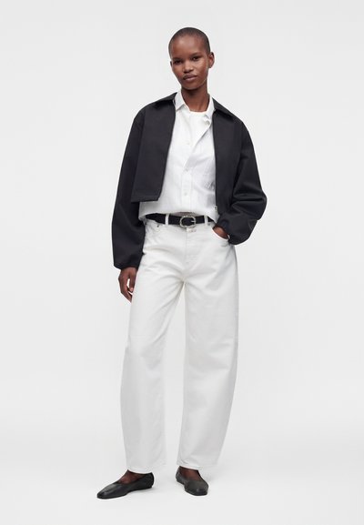 Personne debout avec une main dans la poche, portant une veste noire courte, une chemise blanche boutonnée, un pantalon blanc large, une ceinture noire et des ballerines noires.