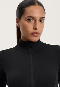 Top sportivo nero con colletto alto e zip intera, realizzato in un tessuto liscio ed elasticizzato senza motivi visibili.