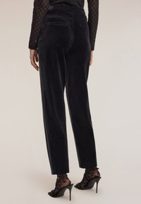 Osoba w czarnych welurowych spodniach z wysokim stanem oraz przezroczystych skarpetach w czarny leopardzi wzór, nosząca czarne szpilki, stoi na jednolitym tle.