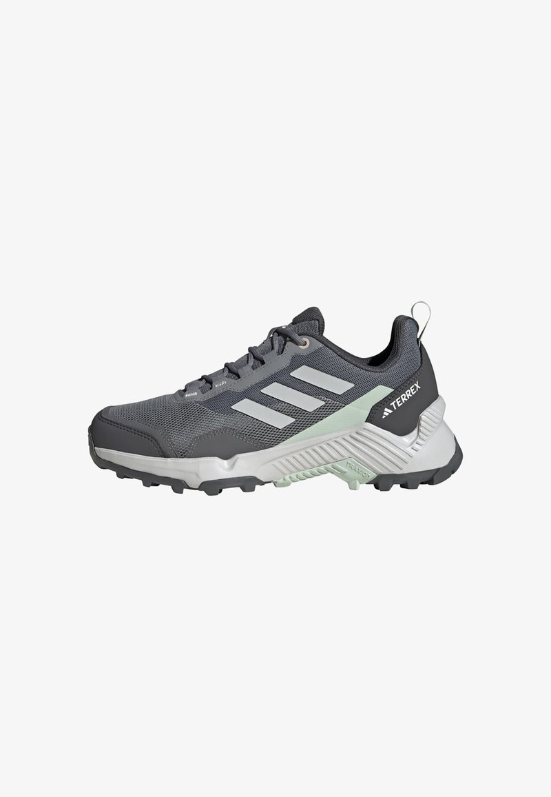Chaussure de sport grise avec une tige en mesh, trois bandes gris clair, un col rembourré et une semelle en caoutchouc noire. Elle présente un embout renforcé et des accents texturés.