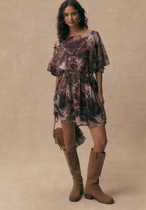 Young woman wearing a floral patterned dress with flutter sleeves, knee-high brown suede boots, and holding a fringed handbag.