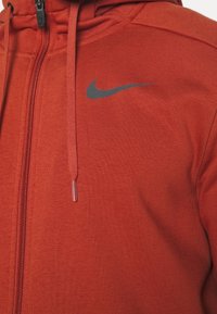 Red zip-up hoodie made of textured fabric, featuring a black Nike logo on the chest and adjustable drawstrings with metallic tips.