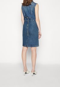 Džínové šaty s fitted siluetou, bez rukávů, s páskem v pase a délku ke kolenům. Mají klasickou modrou barvu a detail ve švu na zadní straně.